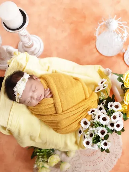 An overhead shot showcasing a beautiful newborn arrangement. The baby is swaddled in a warm yellow wrap, nestled in a basket with coordinating flowers, creating a bright and sunny portrait.