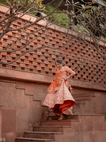 A candid shot of the bride descending the stairs, her orange lehenga in beautiful motion. This photo highlights the grace and elegance of the bridal outfit against the textured architectural backdrop.