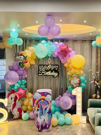 A unicorn and pony themed first birthday party at home. The living room is decorated with a circular balloon arch, character cutouts, and light-up marquee letters for the initial 'S' and number '1'.