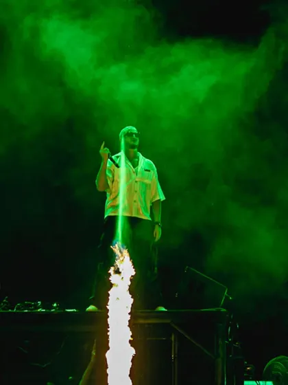 A column of fire shoots up from the stage as DJ Snake commands the crowd, surrounded by green smoke.