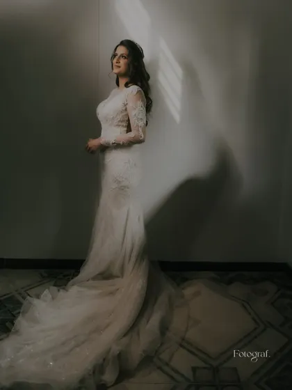 A moody and elegant bridal portrait. The bride stands in a dimly lit room, with a sliver of light illuminating her white lace gown.