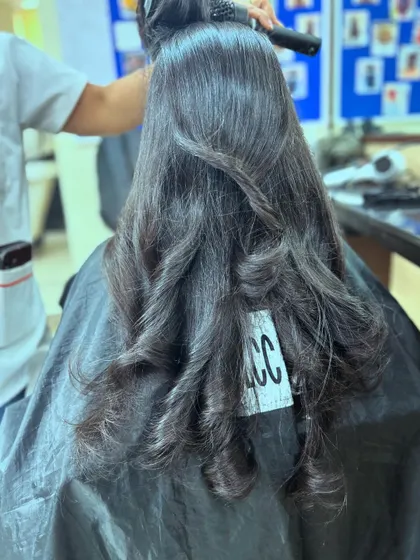 A student practicing her blow-drying technique on a freshly layered haircut. The VLCC-branded cape signifies a professional learning environment.