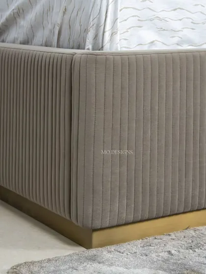 A close-up of the Greco bed's frame. The base is upholstered with vertical baguette stitching and sits on a brushed gold stainless steel skirting, adding a layer of texture and modern luxury.