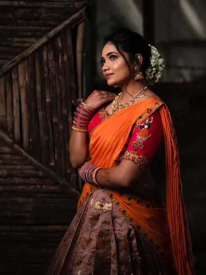 A side profile of the model in the orange and brown half saree, highlighting the hair, makeup, and jewelry that complete the look.