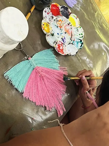 A student adds color to a macrame feather keychain, combining fiber art with painting for a unique, personalized craft.