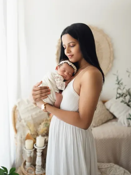 A mother in a beautiful white dress holds her sleeping newborn daughter. The soft, natural light highlights the peacefulness of this moment.