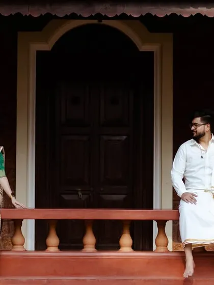 A wide shot of a couple in traditional South Indian attire, sitting on the railing of a veranda of a classic home. This photo uses the location's architecture to frame the couple and tell a story.