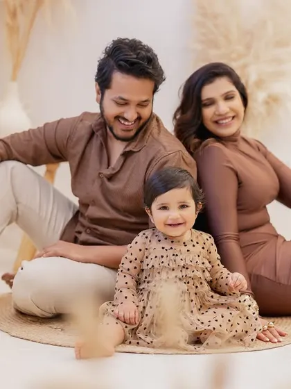 A happy family portrait celebrating a first birthday. The family of three sits together on the floor, with the birthday girl in the center smiling brightly for the camera.