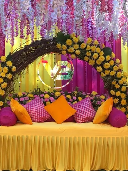 A vibrant seating arrangement for a Haldi or Mehendi function. The backdrop combines bright pink and yellow drapes with a rustic twig arch adorned with yellow marigolds.