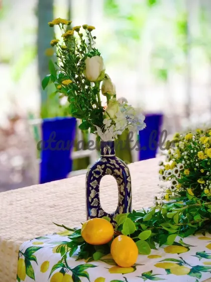 A tablescape detail from a Sicilian-themed event. A blue and white patterned vase with yellow and white flowers sits on a lemon-print table runner, creating a fresh and vibrant look.