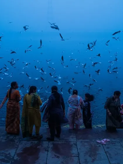 A group of women stand at the edge of the Yamuna river in Delhi, surrounded by a flock of birds in the misty blue morning light.