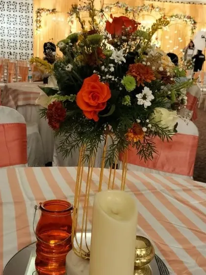 A beautiful tabletop detail from a wedding reception. The centerpiece combines orange roses and other blooms in a modern gold vase, complemented by a large candle for warm lighting.
