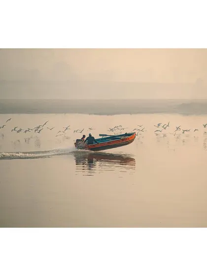 A speedboat cuts across the calm surface of the Yamuna Ghat, with birds taking flight in its wake. The misty background and soft light give the scene an ethereal quality.