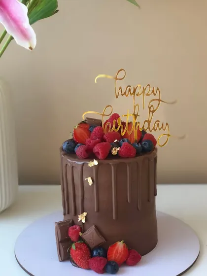A decadent chocolate drip cake topped with a mountain of fresh mixed berries and a gold "Happy Birthday" topper.
