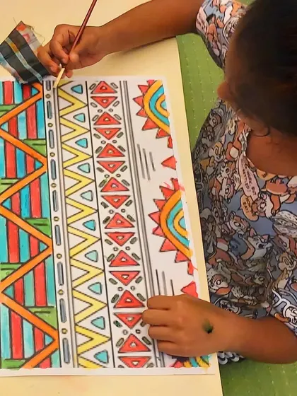 A student works on her vibrant African ethnic pattern painting, carefully filling in the designs with watercolor.