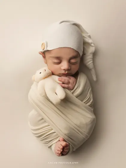 Cute little teddies. This newborn is swaddled in cream with a matching sleepy hat and a tiny bear, creating a dreamy and adorable scene.