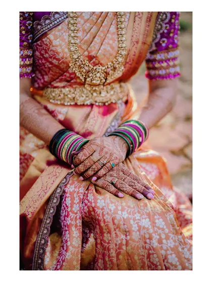 A close-up of the bride's hands, showing the intricate details of her saree and the heavy embroidery on her blouse sleeves.