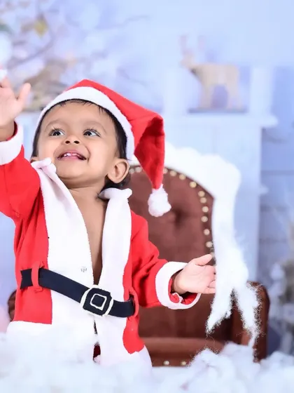 A baby dressed as Santa Claus, waving happily amidst a snowy setup.