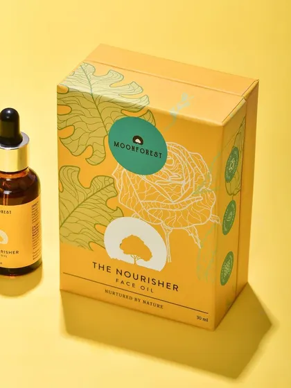 A bright and cheerful shot of Moon Forest's "The Nourisher" face oil next to its box against a yellow background. This is a great e-commerce shot that shows both the product and its packaging.