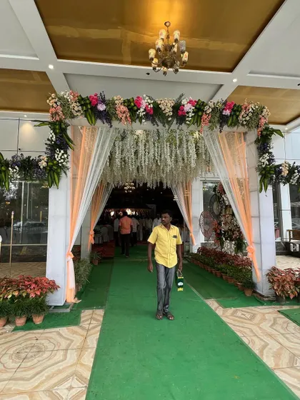 A lovely floral entrance arch for a wedding hall. The combination of pastel-colored flowers, hanging white floral strings, and soft peach drapes creates an elegant and inviting look.