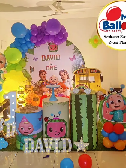 A Cocomelon-themed first birthday with a rainbow balloon arch. The setup includes the iconic yellow bus, character cutouts, and a 'DAVID' marquee.