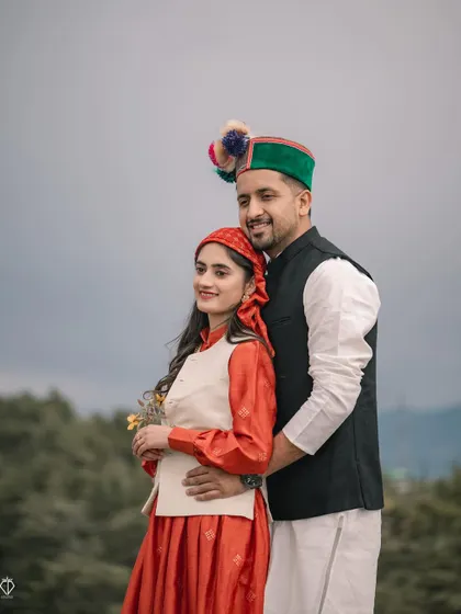 A classic portrait of the couple in full traditional Pahari dress, including the iconic Himachali cap. They stand together against a backdrop of green hills.