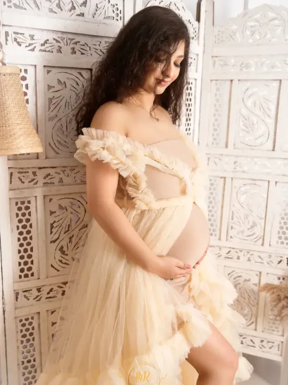 A gentle and serene portrait of an expectant mother. She is wearing a lovely off-the-shoulder cream gown, standing against a white carved screen for a touch of elegance.