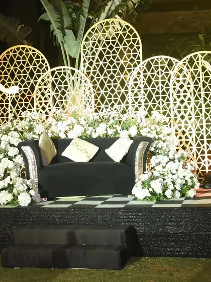 A chic and modern photo booth for a glamorous Sangeet. The setup features a black velvet couch, a backdrop of white geometric latticework, and an abundance of white florals, creating a perfect spot for stylish photos.