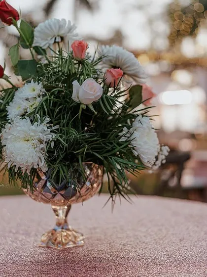 A close-up of a table centerpiece at an outdoor reception. The simple arrangement of roses and chrysanthemums in a crystal vase adds a touch of elegance to the decor.