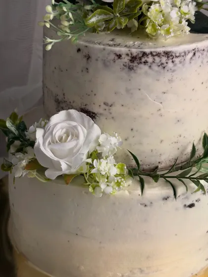 A detailed close-up of my rustic semi-naked cake. You can see the texture of the light buttercream coating over the dark cake, complemented by the delicate arrangement of food-safe white flowers and greenery.