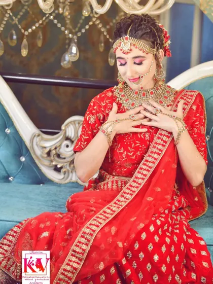 A full-length view of the bride seated, showcasing the elegant red lehenga and the complete set of bridal jewellery. The classic eye makeup and soft pink lips complete this advanced look.