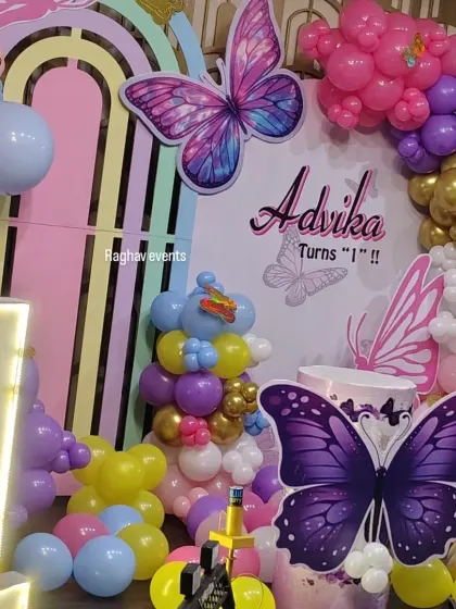 A beautiful butterfly themed first birthday decoration for Advika. The setup includes a pastel rainbow backdrop, a large light-up number '1', and colorful butterfly cutouts.