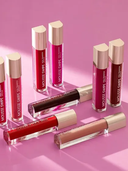 A group shot of PiqueMei's "Mousse Swipe" liquid lipsticks. The products are arranged in a dynamic composition on a solid pink background, with soft shadows adding depth and dimension.