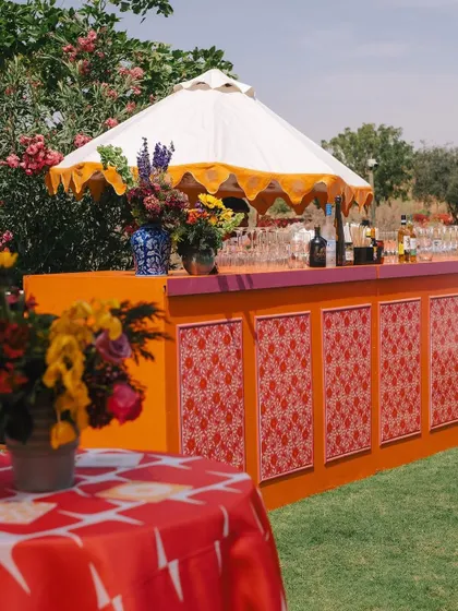 The bar at the Indian bazaar party, decorated with a colorful patterned front and topped with a traditional scalloped umbrella. The floral arrangements add a touch of softness to the vibrant setup.