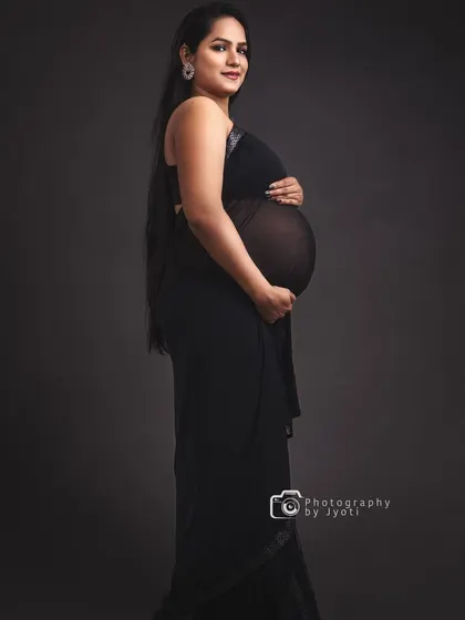 A classic and elegant maternity portrait in a sheer black saree against a dark grey background, highlighting the baby bump silhouette.