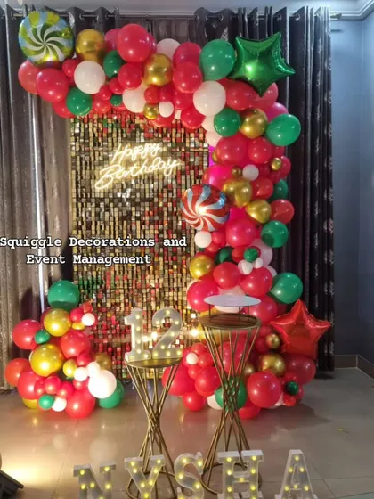 A festive birthday decoration featuring a gold shimmer wall, a 'Happy Birthday' neon sign, and a colorful red, green, and gold balloon arch.