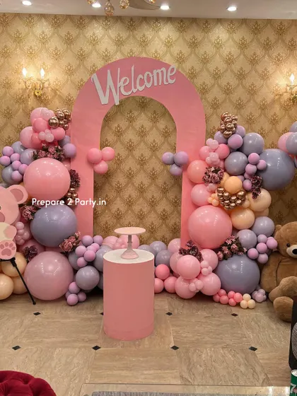 A beautiful welcome party setup featuring a large pink arch with "Welcome" signage. The decor is enriched with a cascade of pink, purple, and rose gold balloons, teddy bear props, and a pedestal for cake.