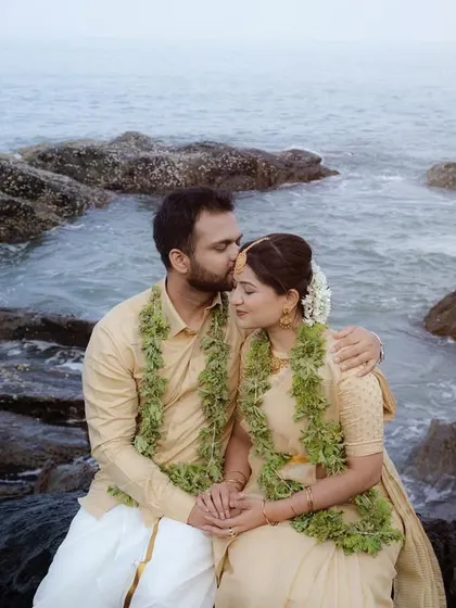 A couple sits on coastal rocks, sharing a quiet moment as the groom kisses the bride's forehead. Dressed in traditional attire with leaf garlands, this image blends romance with a natural, serene beach setting.