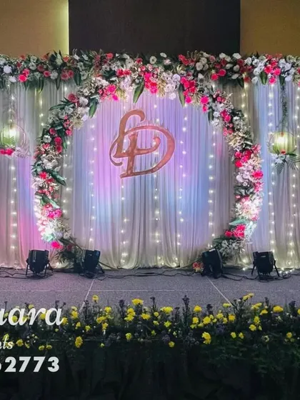 A beautiful stage design featuring a floral ring with a custom monogram, set against a backdrop of fairy lights. The hanging floral lanterns add a whimsical touch to this engagement or reception setup.