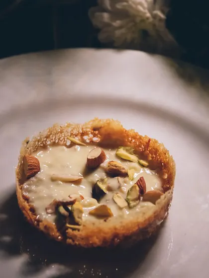 A close-up of a Ghevar tart, a crispy disc topped with creamy rabri and nuts.