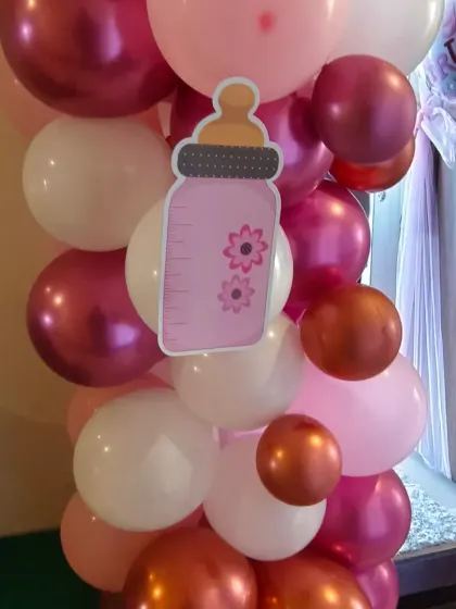 A close-up of a balloon pillar for a baby girl's welcome. The combination of chrome, metallic, and pastel pink balloons with a cute baby bottle cutout shows attention to detail.