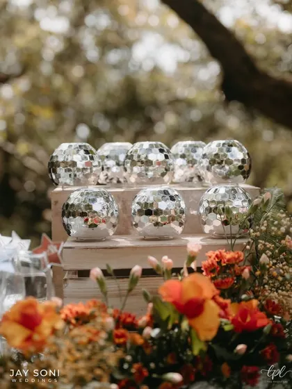 Small disco ball candle holders add a touch of sparkle to a rustic wooden crate, set against a backdrop of vibrant orange flowers.