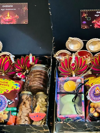 A pair of black marble-patterned boxes filled with Diwali essentials like diyas, lotus-shaped decor, snacks, and candles.