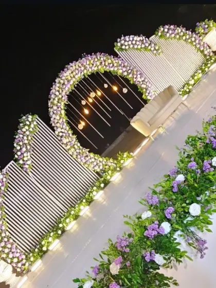 This modern stage design uses a unique combination of textures and shapes. A circular floral arch is set against a backdrop of white panels, with hanging lights that create a waterfall effect. The purple and white flowers add a touch of soft color to the contemporary structure.
