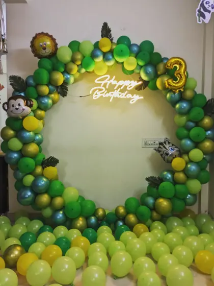 A simple yet fun jungle theme birthday decoration. A circular balloon arch in shades of green is accented with monkey and lion foil balloons and a neon sign.