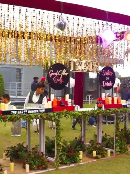 A lively outdoor bar setup for a day wedding. The bar is decorated with greenery and candles, with fun signs and a canopy of gold tassels to create a festive vibe.