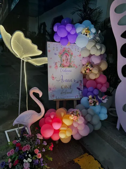 A welcome sign for "Avana's One-derland" decorated with a colorful balloon garland and flanked by a flamingo and an illuminated butterfly prop.