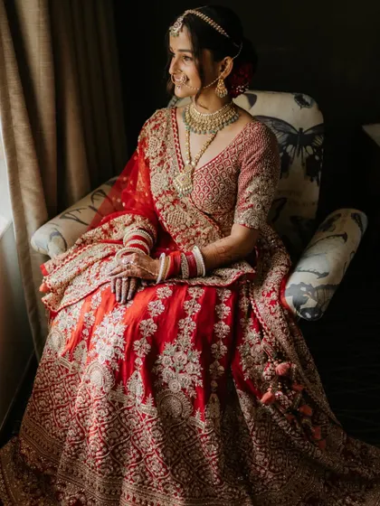 A classic bridal portrait of the bride seated, her stunning red lehenga beautifully arranged. The soft window light highlights her grace and elegance.