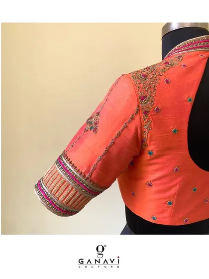 A vibrant coral-pink blouse with a high neck and detailed embroidery on the sleeves. The craftsmanship showcases a blend of different threads and techniques to create a rich, textured cuff.