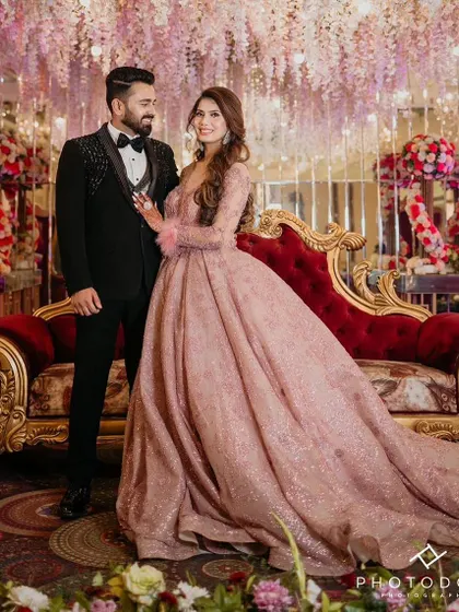 Our client and her partner looking picture-perfect on their engagement day. She is wearing our dusty pink long-sleeved ball gown with feather cuffs, which looks absolutely stunning against the floral backdrop. A true princess moment.